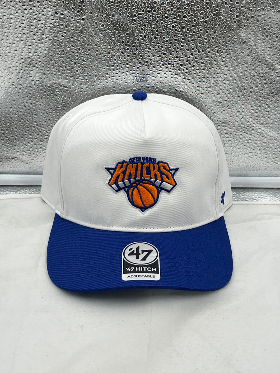 New York Knicks NBA '47 Brand White Two Tone Hitch Adjustable Snapback Hat displayed on a wooden table with a blurred basketball background.